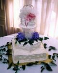 Three layer white cake.  Square base cake with hexagonal cake atop the base.  Then clear pillars holding up a round top cake.  Purple flowers on top of 2nd layer, crown  with gauze and pink ribbons on top layer