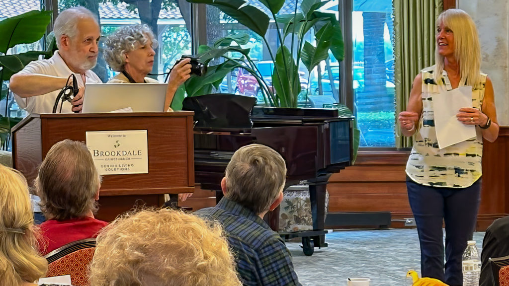 Bernie at a podium with a microphone and computer, woman with a camera, and a woman holding a song sheet