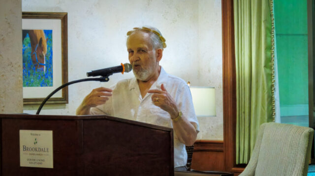 Man at podium speaking into microphone, gesturing while wearing a gold laurel leaf tiara