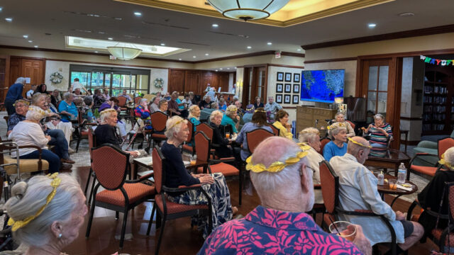Large group of seated people, wearing gold laurel leaf tiaras, listening to the host who is off camera to the right