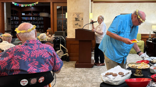 Refreshment table including Mayoff Day chocolate peanut butter cookies and man serving himself. Man in background at podium speaking into microphone